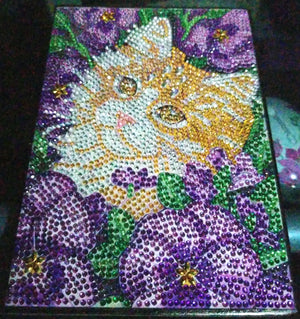 Notesbog med kat omgivet af lilla blomster. Diamond paint.
